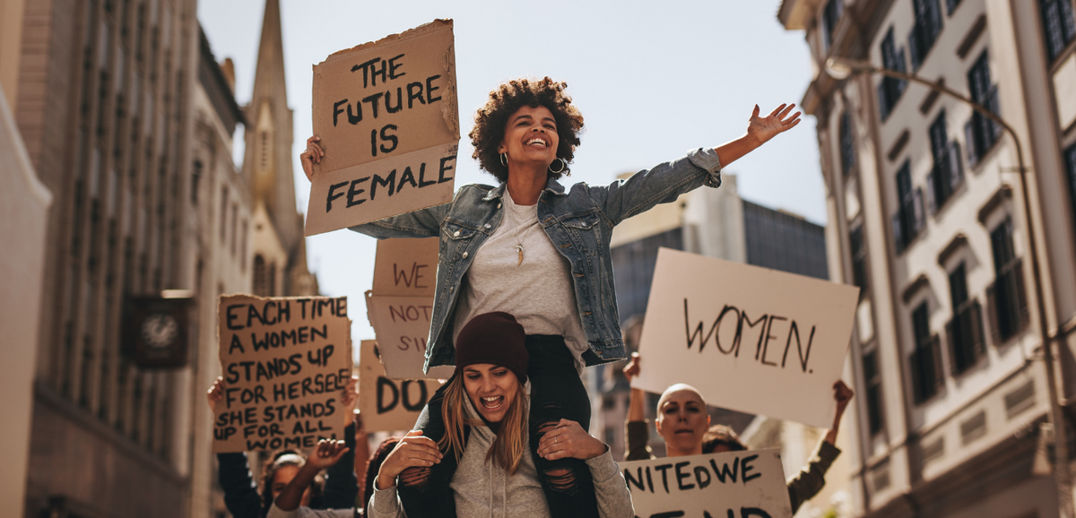 A woman on another woman's shoulders at a protest, holding a sign that says 'The Future is Female' and surrounded by others with signs that say 'Women' and 'Each time a woman stands up for herself, she stands up for all women.'