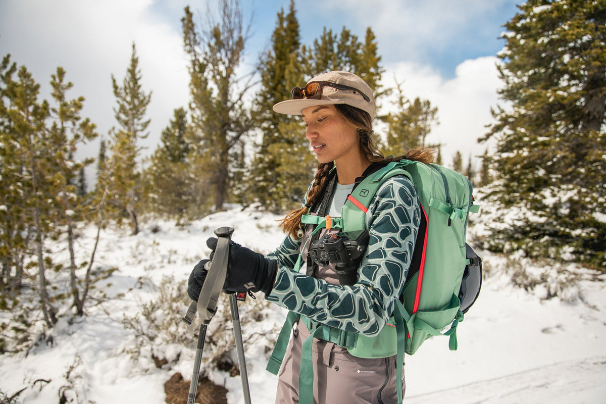 A woman in a green backpack and patterned shirt, holding ski poles in a snowy landscape.