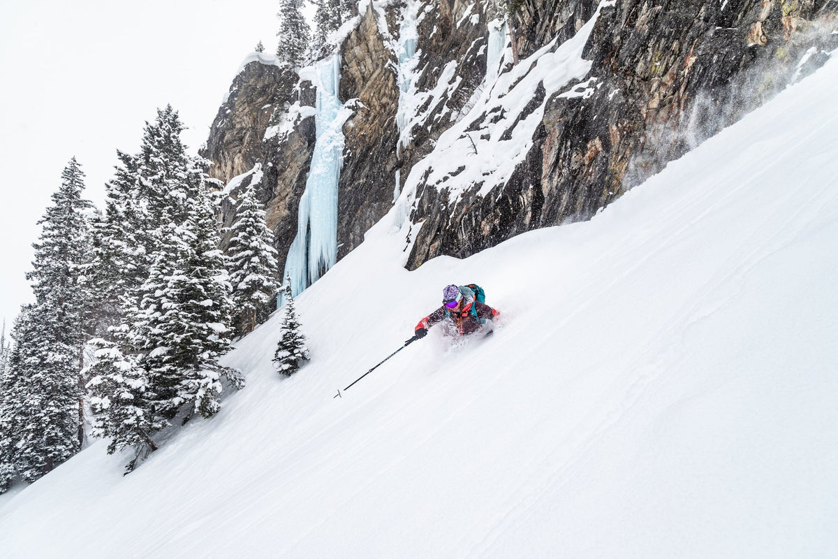 A female skier navigating through deep snow with a waterfall in the background.
