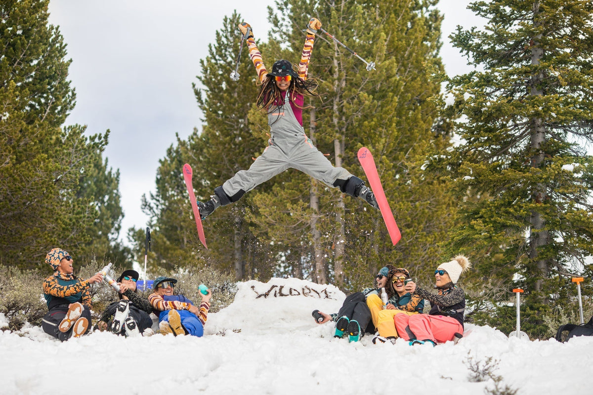A skier jumping in the air with a group of people sitting in the snow, surrounded by trees.