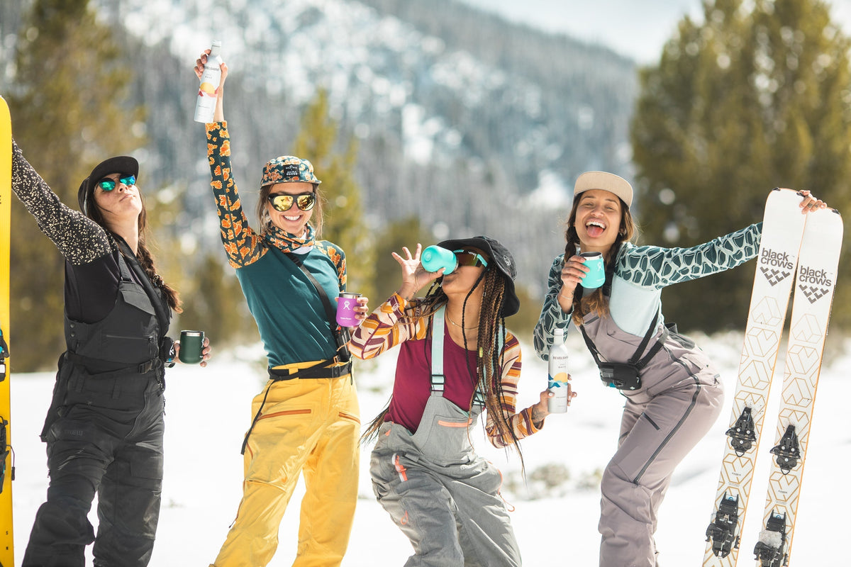 Four women celebrating in winter gear, holding drinks and ski equipment in a snowy landscape.