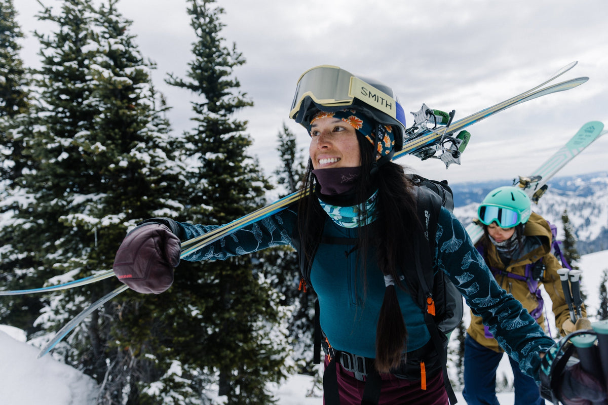 A woman smiling while skiing uphill with another skier in the background, surrounded by snow-covered trees.