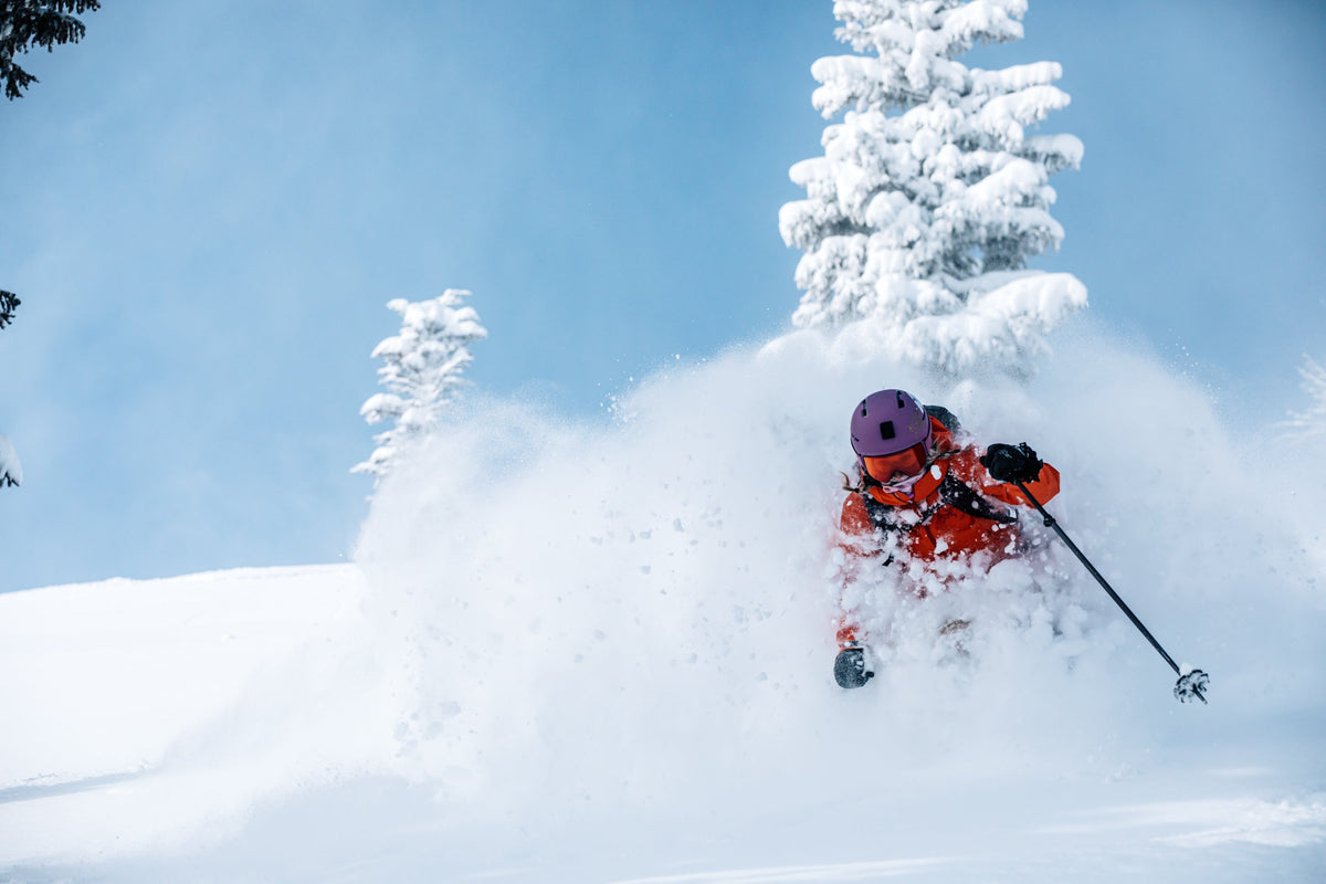 A skier in an orange outfit skiing through deep snow with trees in the background.