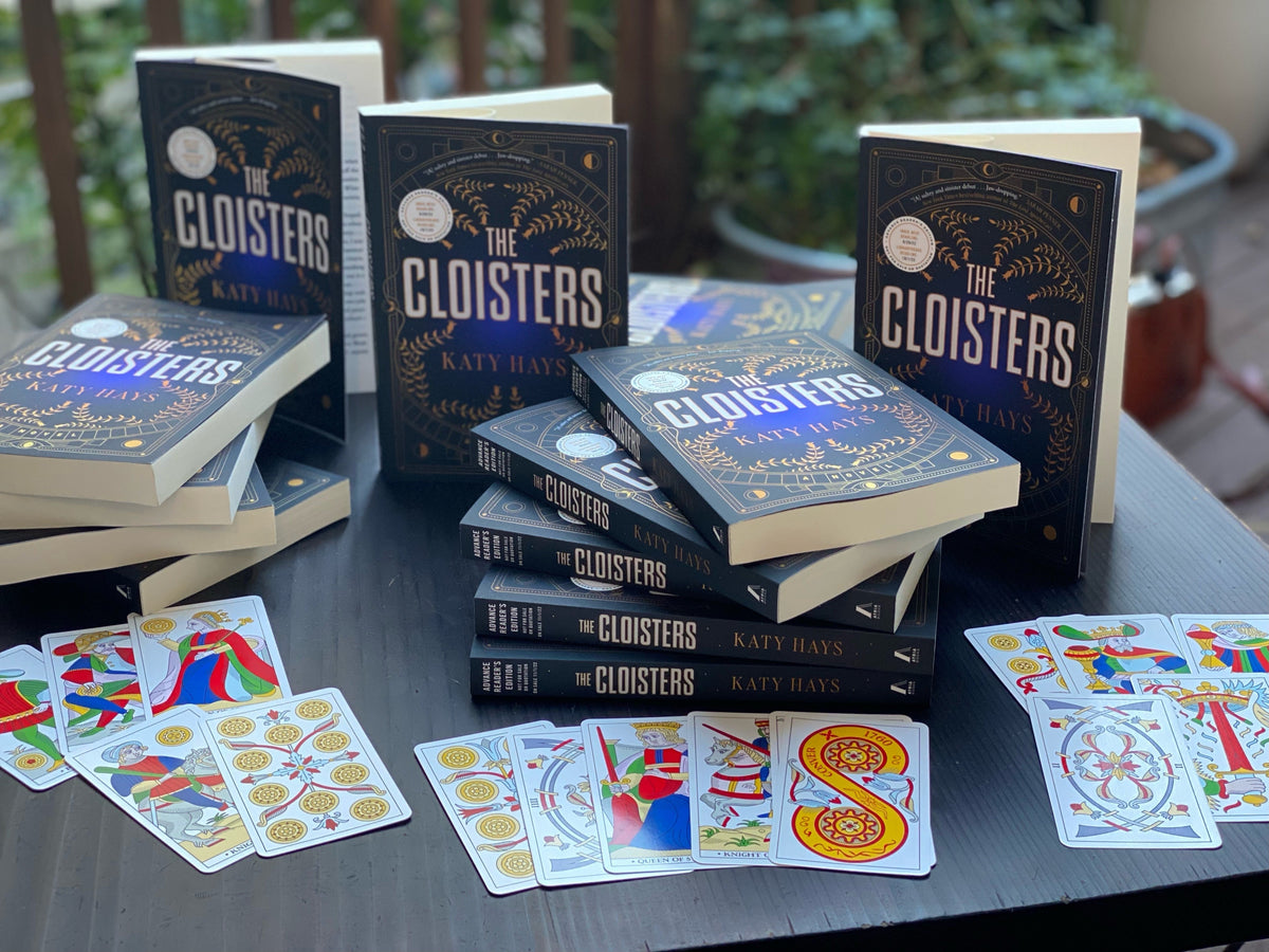 A stack of books titled 'The Cloisters' by Katy Hays with tarot cards on a table.