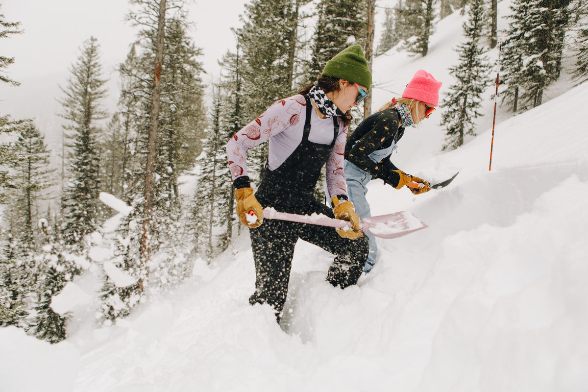 Two women shoveling snow in a winter landscape, wearing colorful winter gear.
