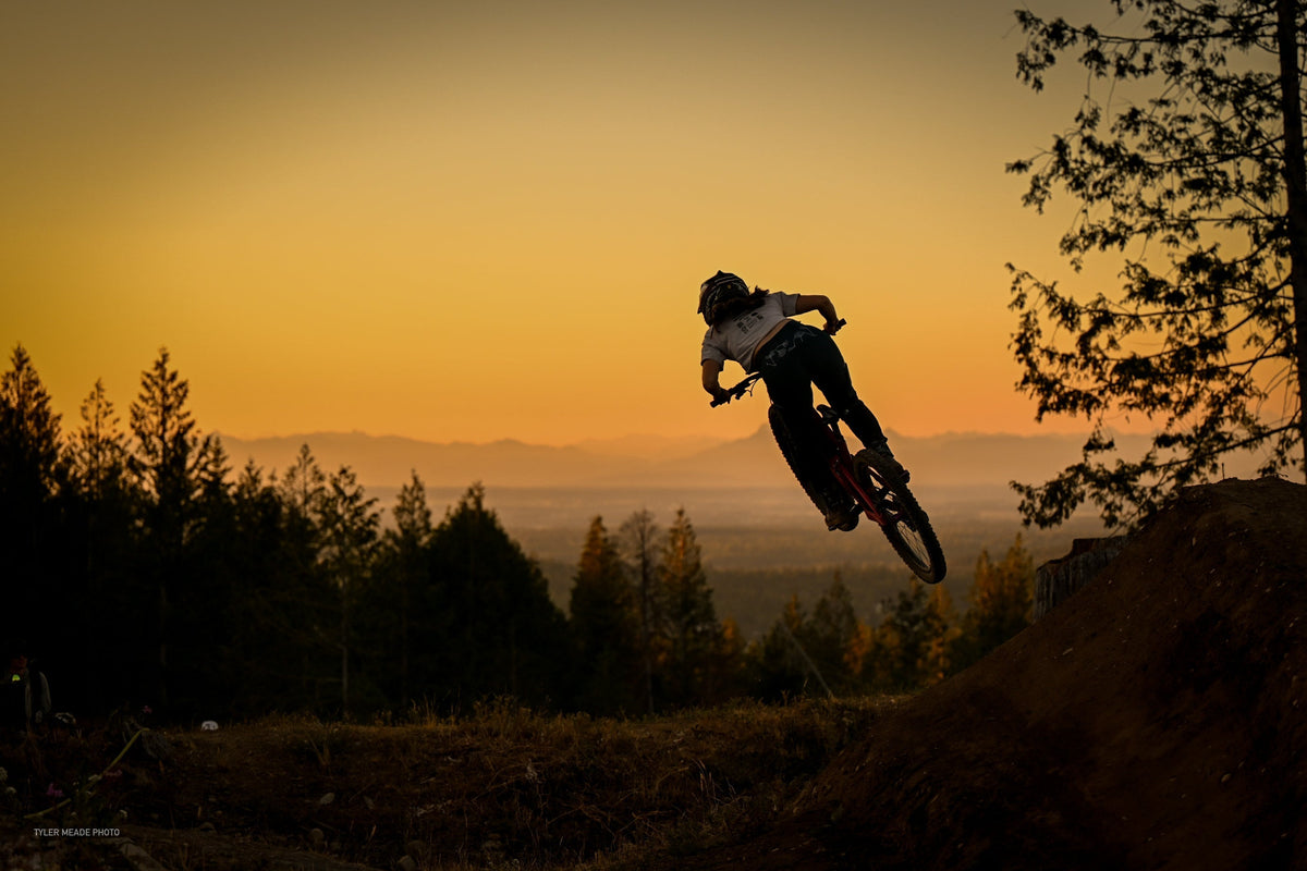 A mountain biker jumping against a sunset backdrop with trees in the foreground.