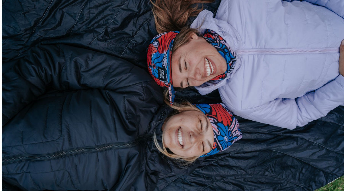 Two women smiling while wearing jackets from the Payette collection, lying on a black surface.