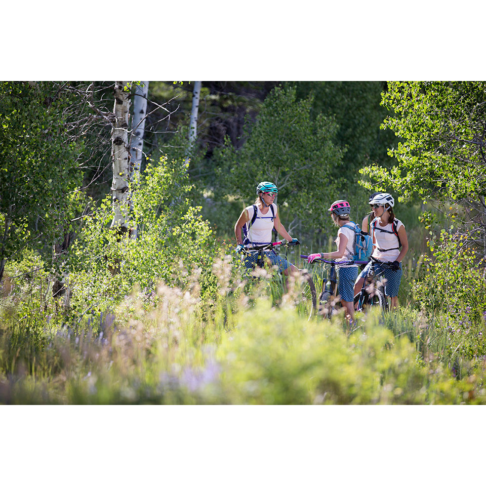 Three women in biking gear discussing on a trail surrounded by greenery