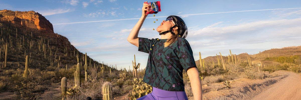 Two women outdoors on a dirt trail wearing women's bike tops; one in a light purple top with a small black logo on the back, the other in a dark green top with purple and pink pants.