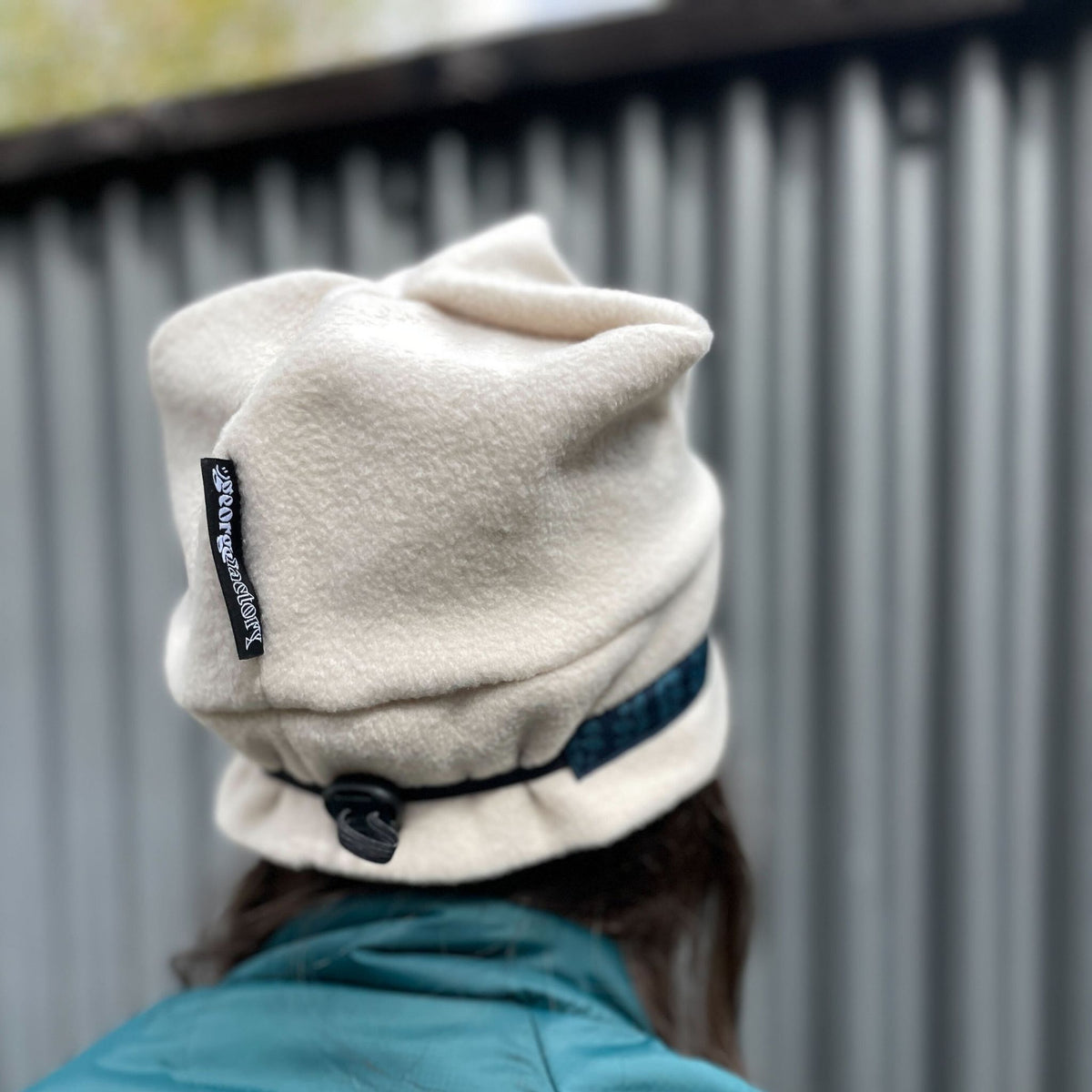 Person wearing a white beanie with a logo and a blue jacket, standing against a metallic wall.