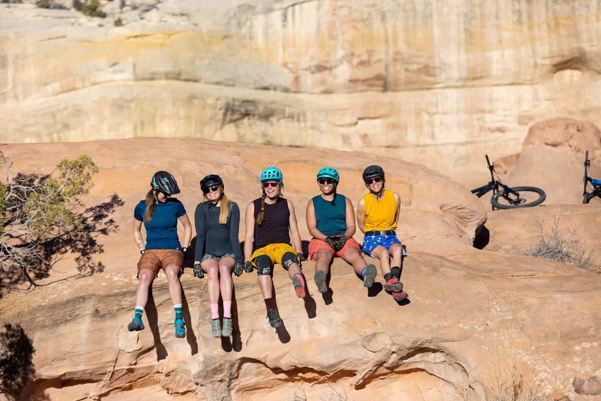 Five women in cycling gear sitting on a rock formation, with bikes in the background.