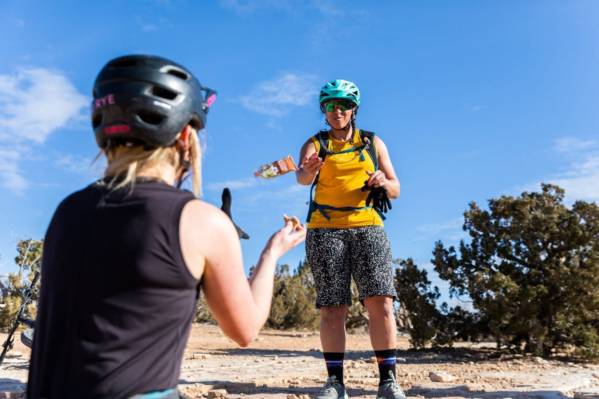 Two mountain bikers sharing snacks outdoors, one wearing a helmet and yellow shirt.