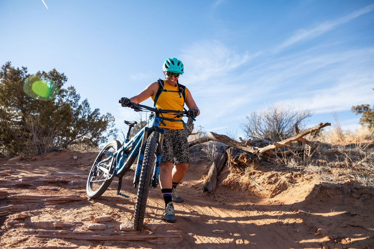 A woman in a yellow shirt and helmet walking a mountain bike on a trail