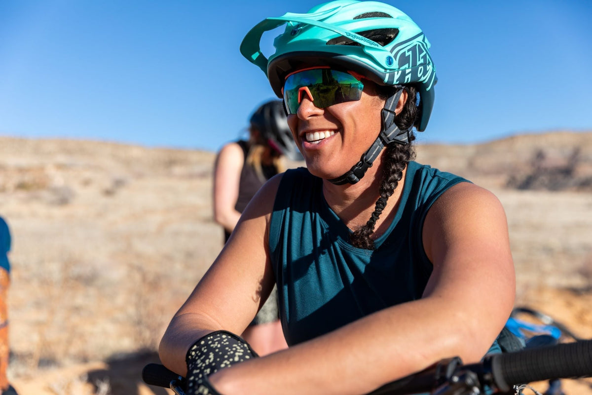 A smiling woman in a teal helmet and sunglasses, enjoying a biking adventure in a desert landscape.
