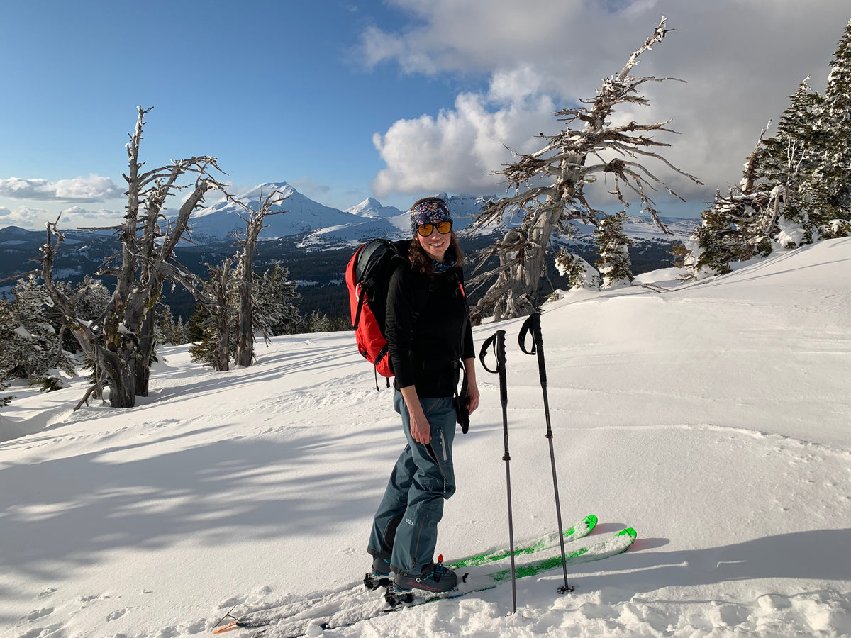 A skier standing in a snowy landscape with mountains in the background, wearing a backpack and ski gear.