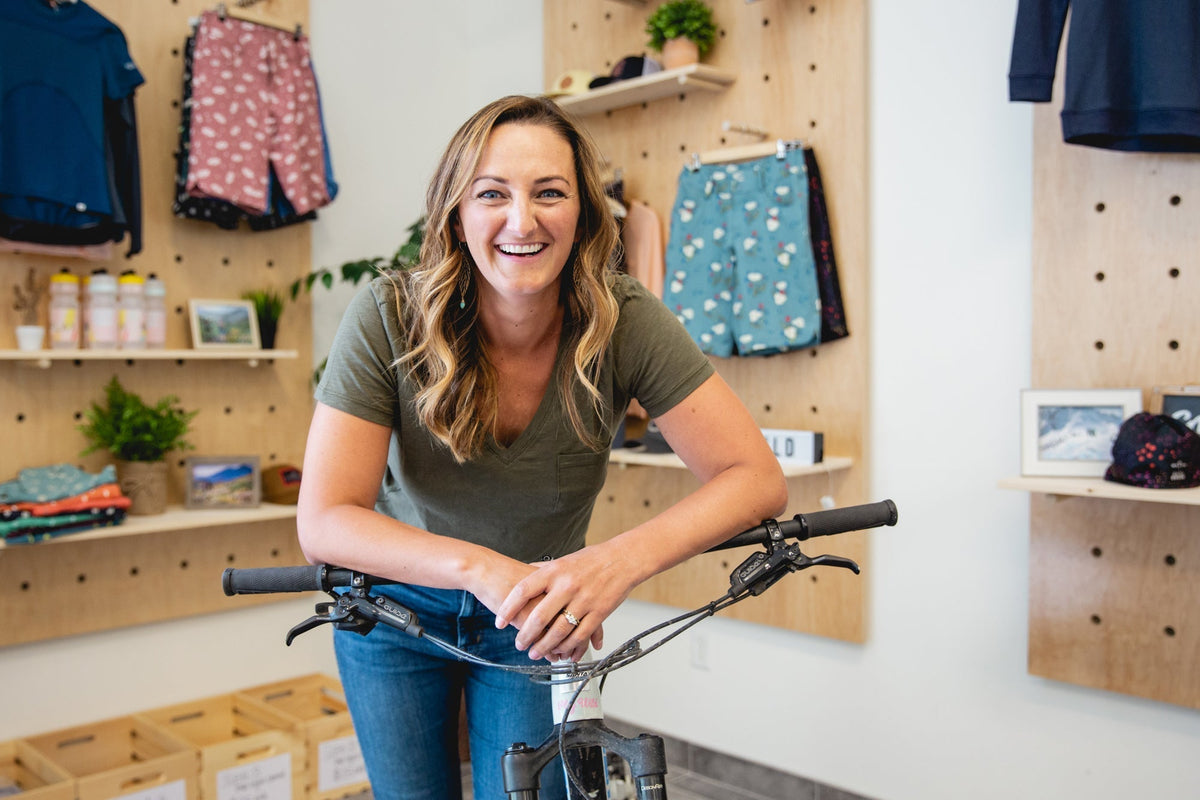 Smiling woman in a green shirt leaning on a bicycle in a clothing store with hanging garments.