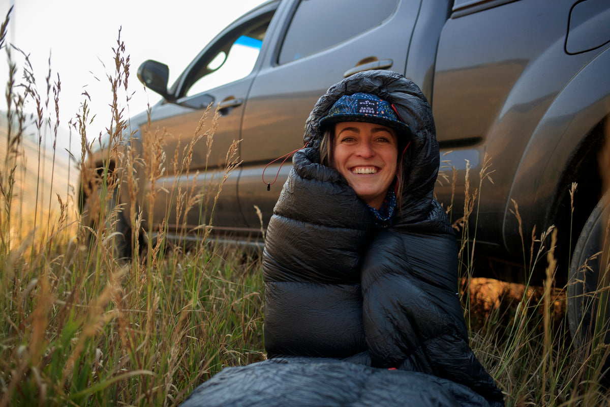 A smiling woman wrapped in a black puffy sleeping bag, sitting in grass near a vehicle.