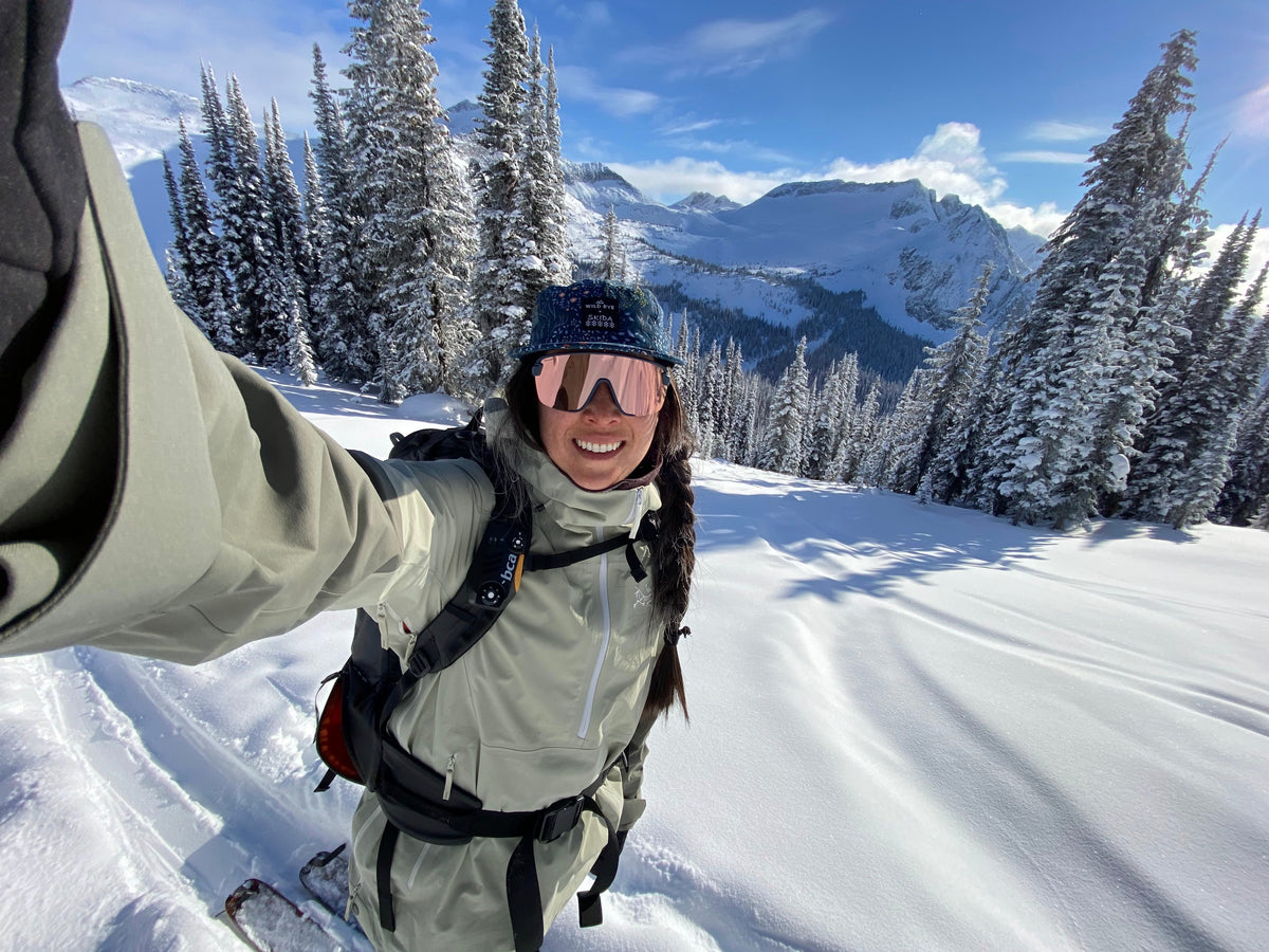 A person in winter gear smiling in a snowy landscape with mountains in the background.
