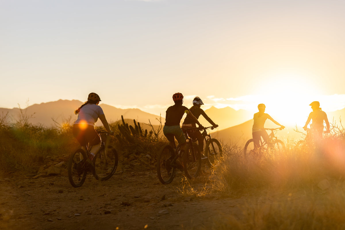 Five cyclists riding on a dirt trail during sunset with mountains in the background.