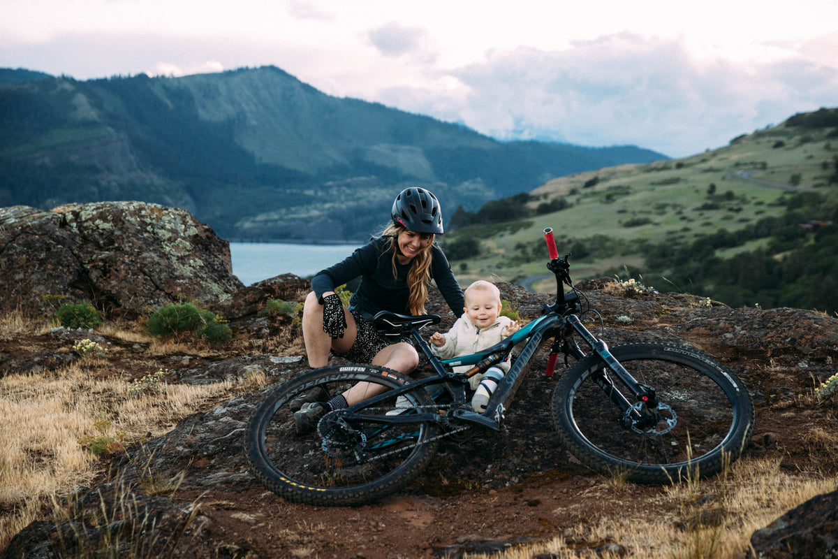 A woman with a helmet and a child beside a mountain bike in a scenic landscape.
