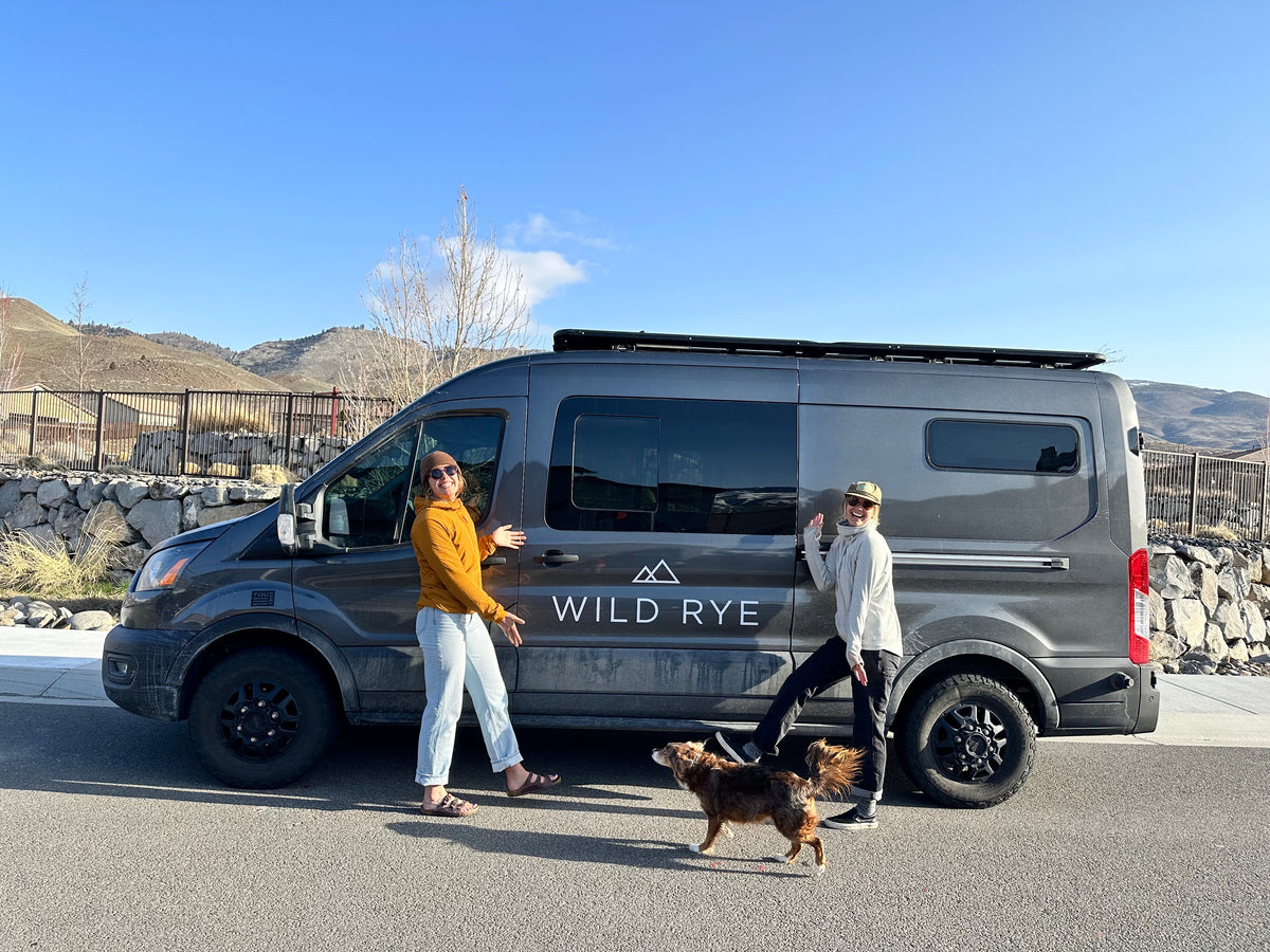 Two women and a dog next to a 'WILD RYE' branded van in a sunny outdoor setting.