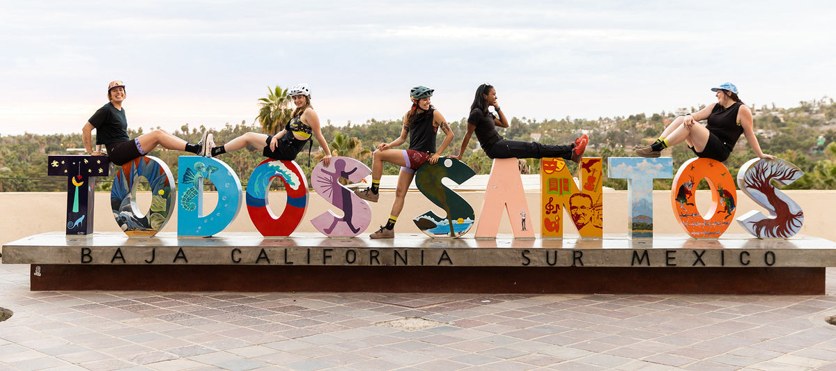 Group of women posing by a colorful sign that reads 'TODOS SANTOS' in Baja California, Mexico.