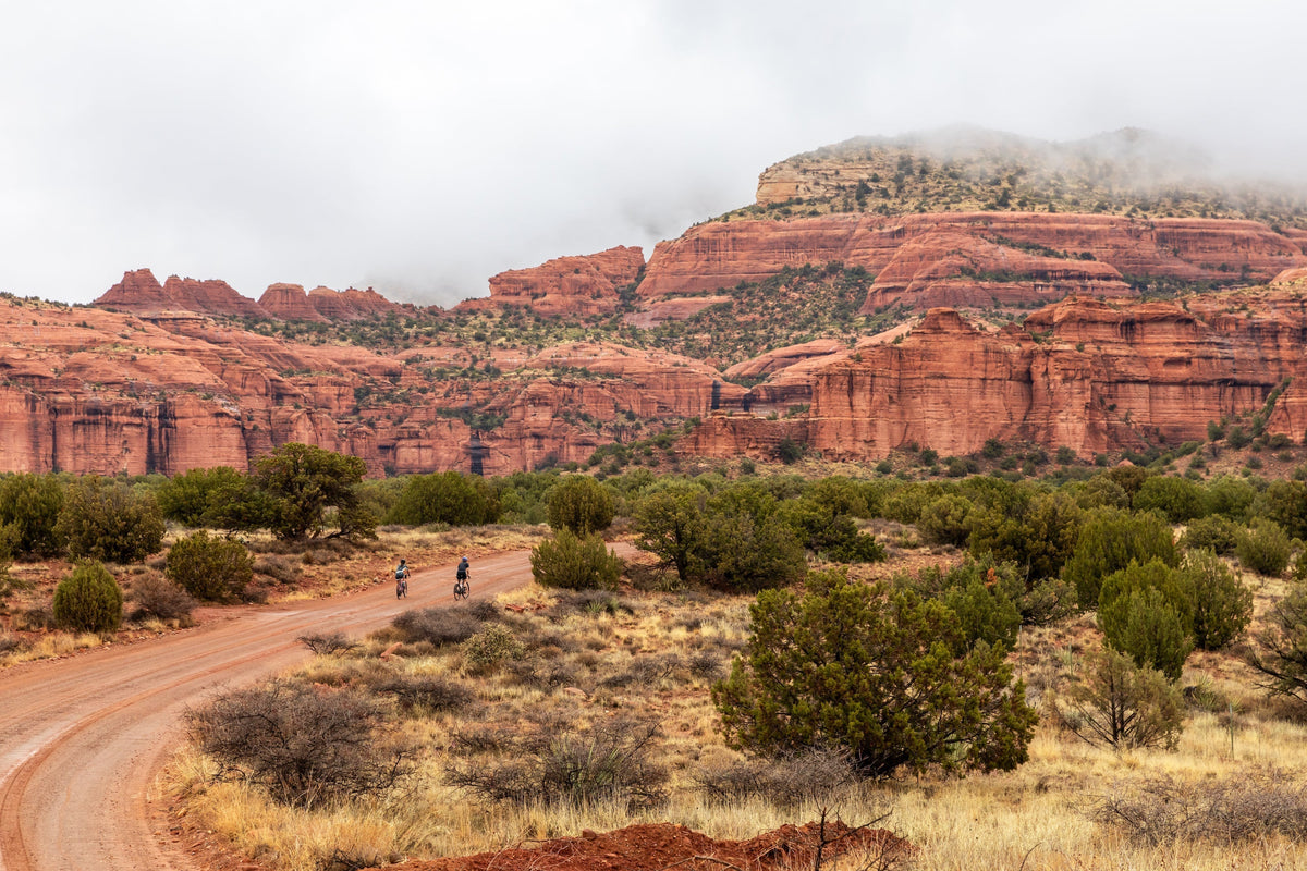 Two mountain bikers riding on a dirt road with red rock formations in the background