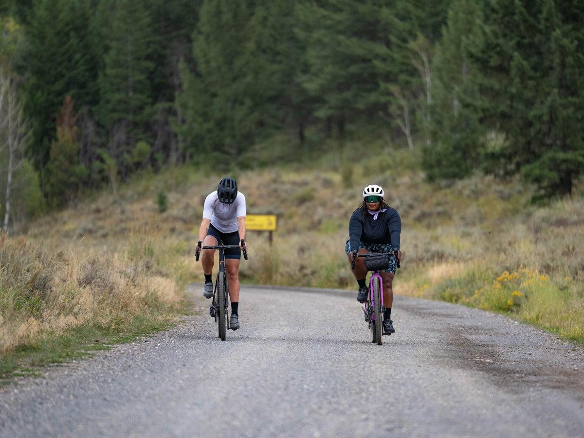 Two cyclists riding on a gravel road surrounded by trees.