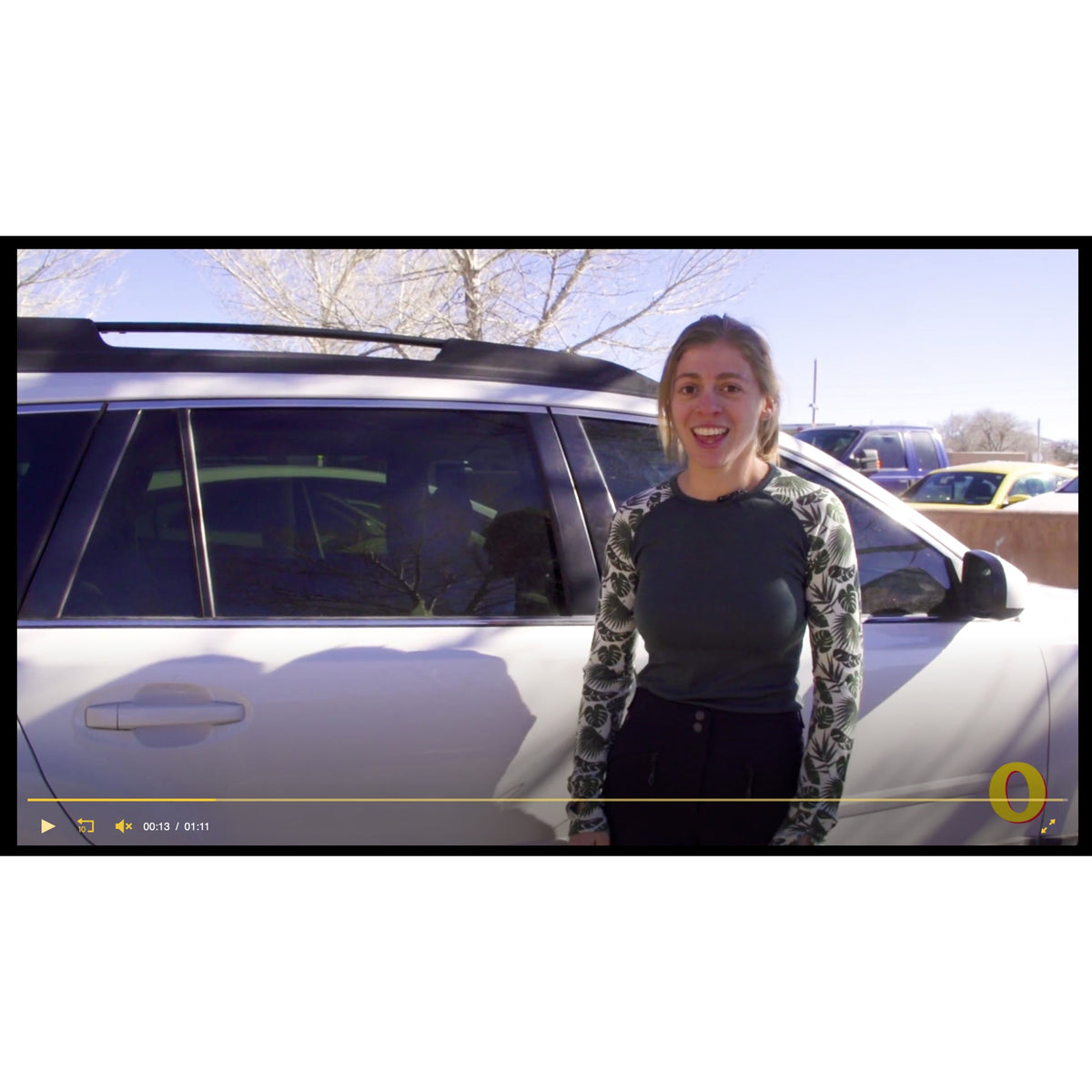 A woman wearing a floral long-sleeve shirt stands next to a white SUV in a parking lot.