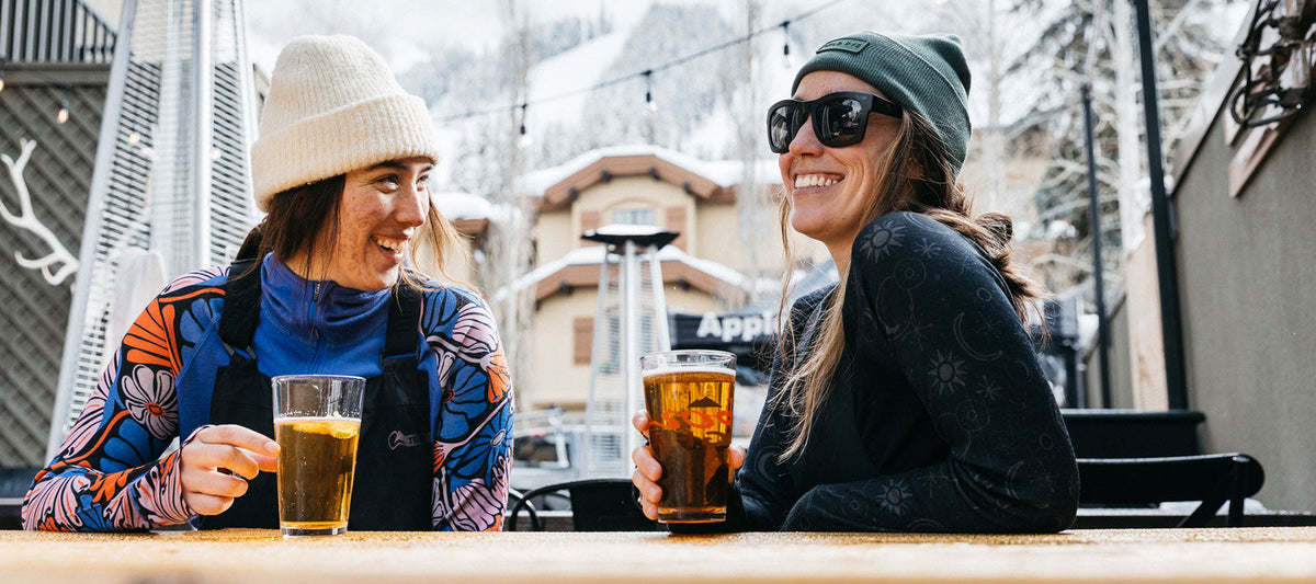 Two women wearing colorful 100% merino baselayers and winter hats, sitting outdoors at a table with drinks, smiling and talking, with snowy mountains in the background.