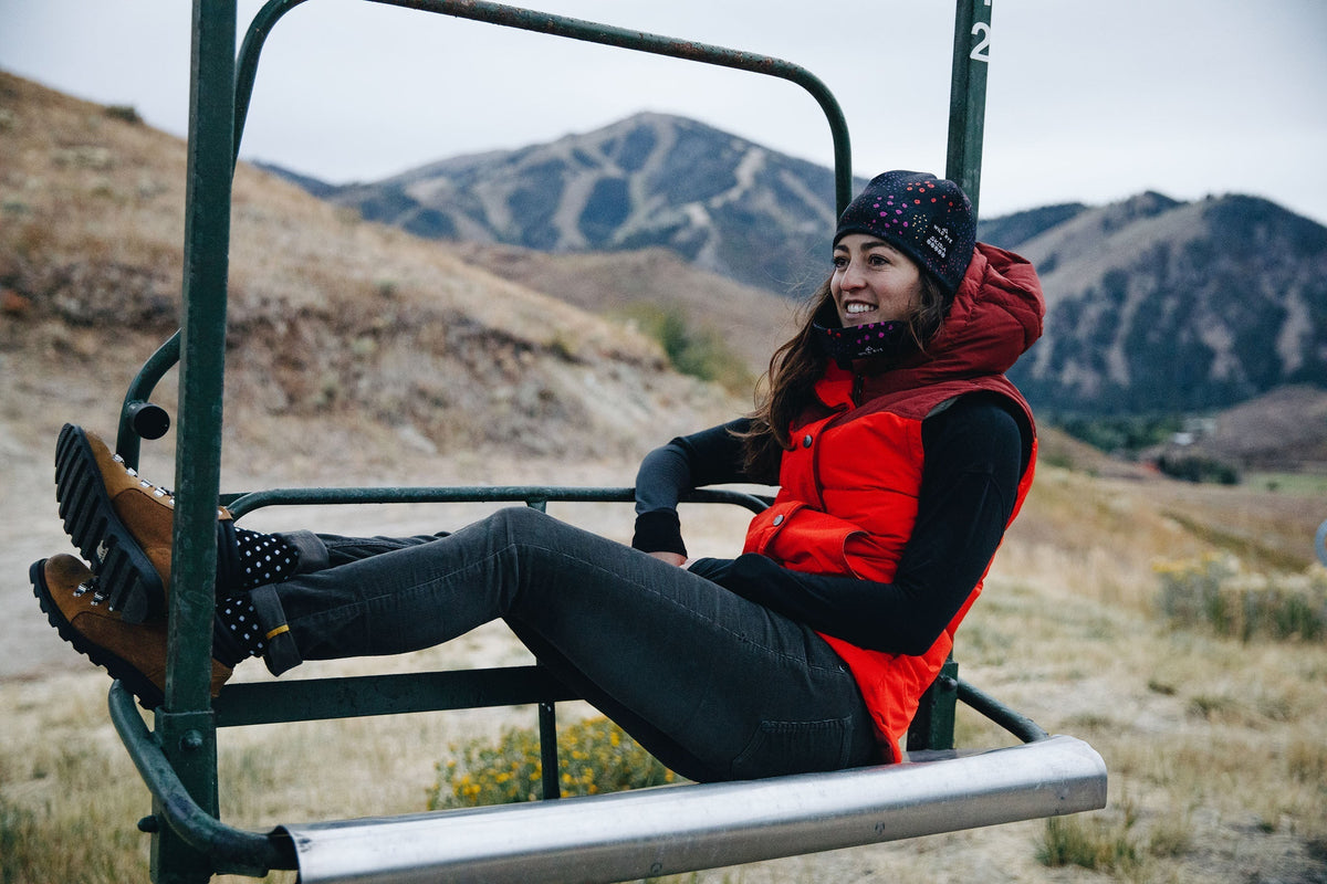 Woman wearing a red vest, black long sleeve shirt, black beanie with pink dots, and brown boots sitting on a ski lift chair with mountains in the background.