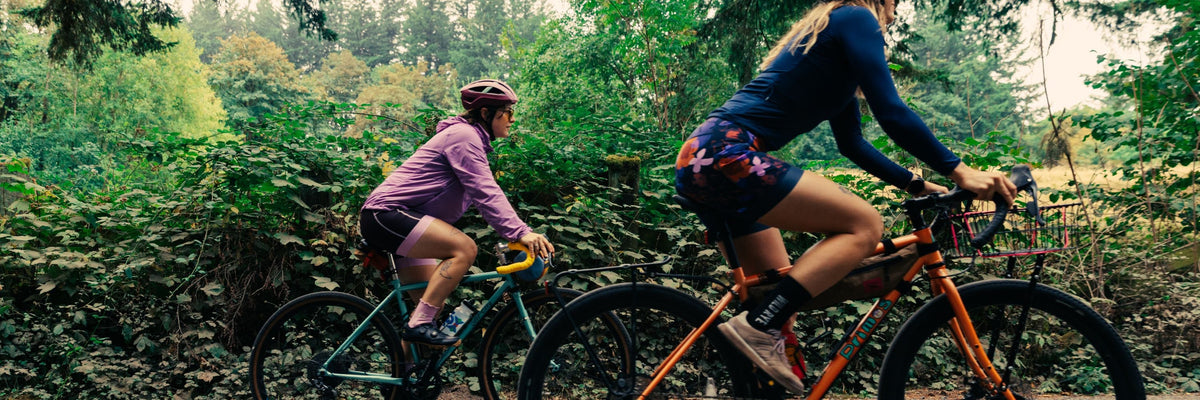 Cyclist wearing a pink top and floral shorts riding a black bicycle on a forest trail, motion blur in background