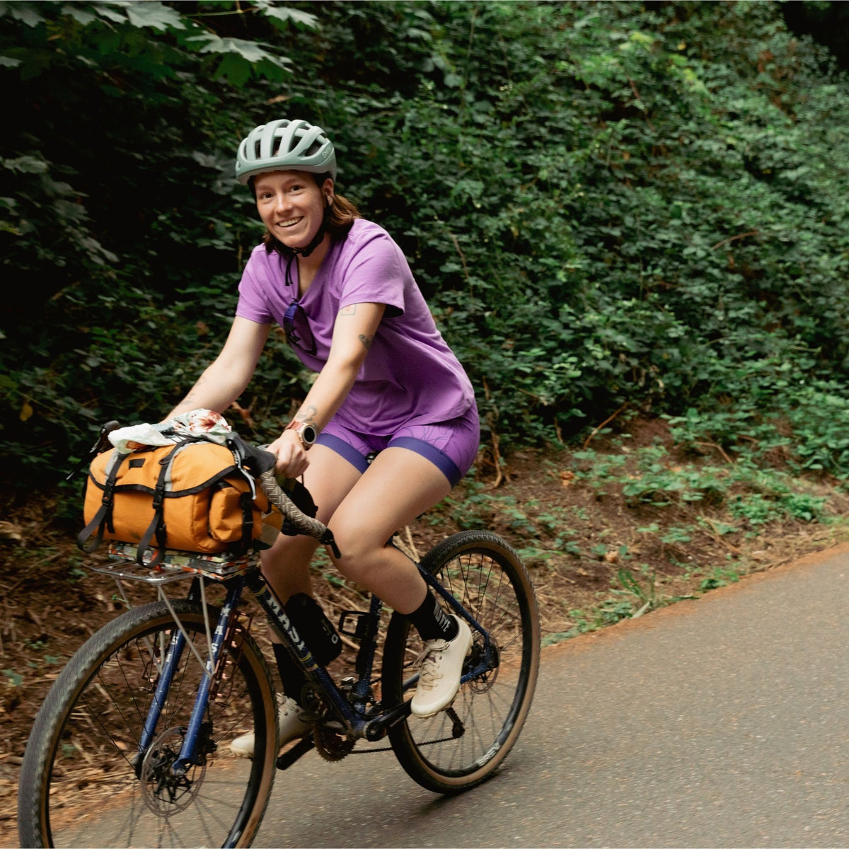 Woman riding a bicycle on a trail with greenery in the background wearing purple shirt and shorts