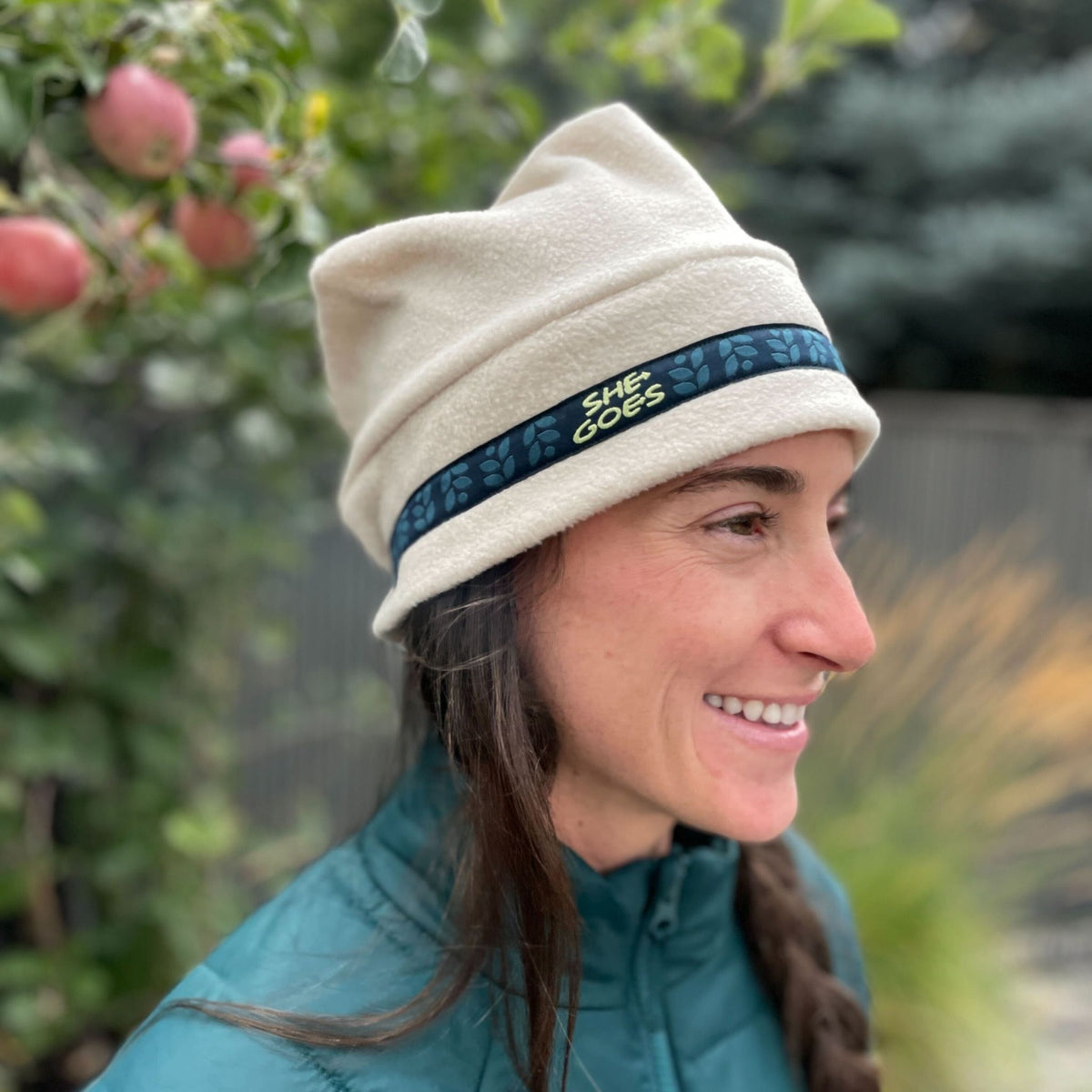 Woman wearing a beige hat with a blue and green band featuring 'SHE GOES' text, standing outdoors with greenery and apples in the background.