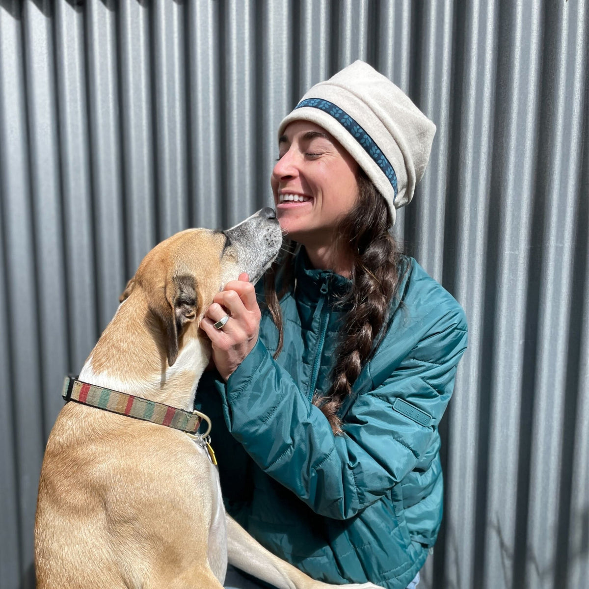 Woman in a green jacket and white beanie interacting with a dog against a corrugated metal background