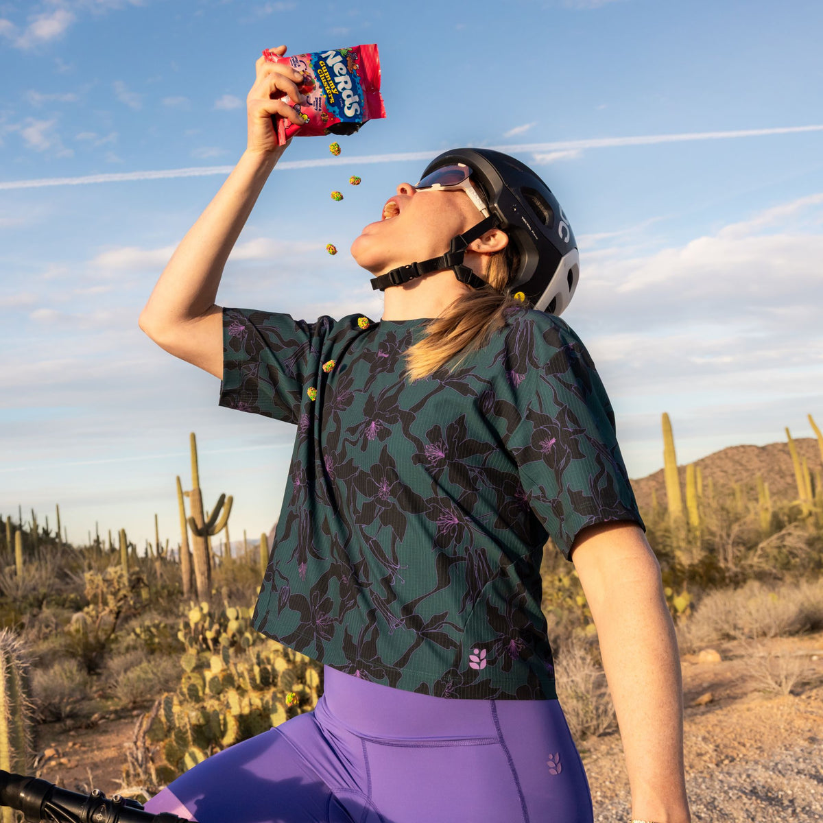 [Spruce Aquila Bloom] Person in a desert setting eating nerds clusters, with cacti and mountains in the background.