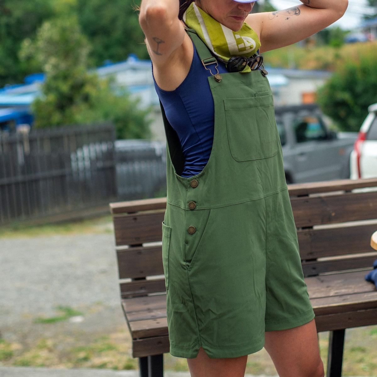 [Olive You] Person wearing green overalls standing near a wooden bench outdoors.