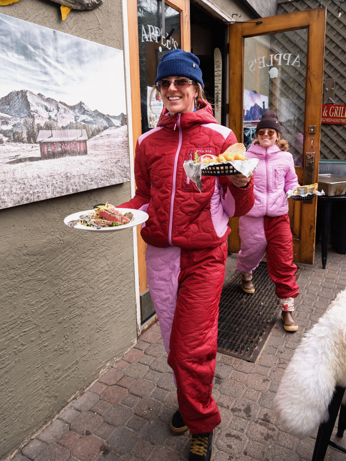 [Ruby Blocked] Two people wearing matching red and pink payette outfits walk out of a shop doorway holding food trays 
